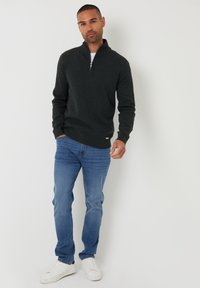 Donkergrijze sweater met een half-rits kraag, gedragen over een wit shirt. Nokte met lichtblauwe jeans en witte sneakers. Vlotte textuur.