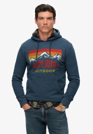 Marineblauwe hoodie met berggrafiek, "Superdry Outdoor" tekst in rood en geel, voorzak, gelaagd over een geruit shirt.