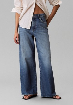 Personne portant un jean bleu pattes d’eph, une chemise beige rayée partiellement sortie et des sandales noires à brides sur fond gris uni.