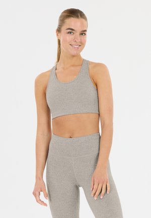 Smilende kvinde med trukket hår tilbage iført lysegrå atletisk crop top og højtaljede leggings mod en ensartet hvid baggrund.