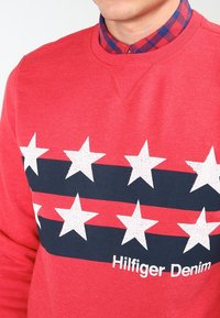 Sudadera roja con una franja azul marino con estrellas blancas agrietadas y el texto "Hilfiger Denim" en la parte inferior. Tela con textura y diseño de cuello redondo.