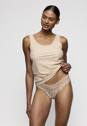Donna con canottiera beige e biancheria intima in pizzo abbinata, che solleva leggermente la canottiera, su uno sfondo semplice di colore grigio chiaro.