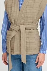 Gilet beige matelassé avec une ceinture nouée, comportant deux poches à l'avant. En dessous, une chemise rayée bleue et blanche est visible.