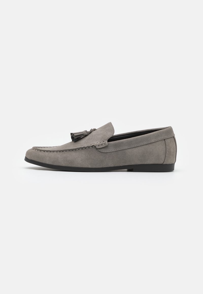 Mocassin pour homme en daim gris avec des glands, semelle noire et détails cousus le long du bord supérieur, présenté de profil sur fond blanc.