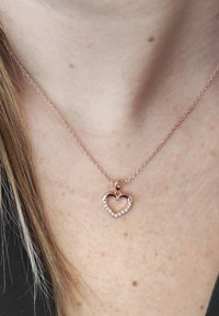 Collana in oro rosa con un pendente a forma di cuore impreziosito da pietre trasparenti, appesa a una delicata catena, poggiata sulla pelle.