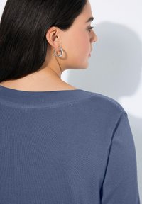 Blauer gestrickter Pullover mit weitem Ausschnitt, der ein strukturiertes, geripptes Muster an den Schultern aufweist. Komplett mit silbernen Creolen.