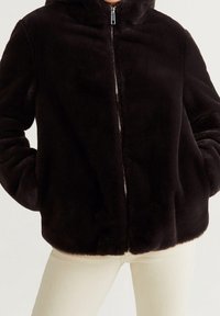 Veste en fausse fourrure marron avec capuche, fermeture éclair et poches latérales. Texture douce et forme ample, idéale pour un style décontracté.