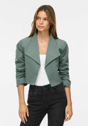 Kvinde med langt brunt hår iført en kort grøn blazer over en hvid top og sorte jeans med høj talje, stående foran en ensfarvet baggrund.