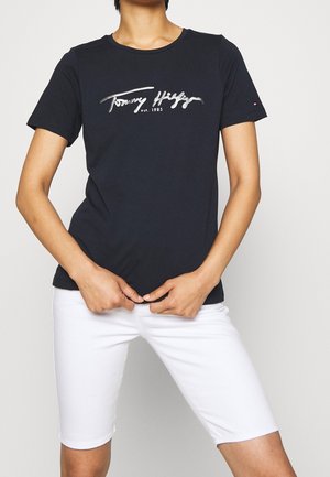 Personne portant un t-shirt noir ajusté Tommy Hilfiger avec un logo blanc en écriture et un short blanc longueur genou, mains ajustant l'ourlet du t-shirt.