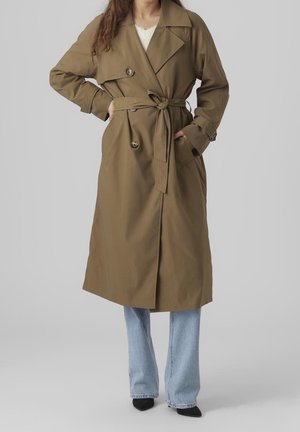 Kobieta w długim brązowym trenczu z paskiem, nałożonym na jasnoniebieskie dżinsy, i czarnych, spiczastych butach, stojąca z rękami w kieszeniach.