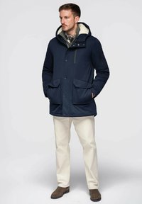 Parka isolée bleu marine avec capuche, doublure en fausse fourrure crème, deux poches avant et poche zippée sur la manche. Associée à un pantalon de couleur claire.