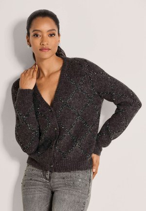 Vrouw met donker haar draagt een donkergrijze gebuttonede cardigan met een ruitpatroon en lichtgrijze versierde spijkerbroek, poserend met één hand op de nek.