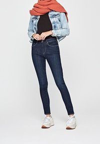 Hubené tmavě modré džíny vyrobené z denimu, kombinované s černým topem a světlou denimovou bundou. Kolem krku je zavěšený texturovaný šátek v barvě rzi.