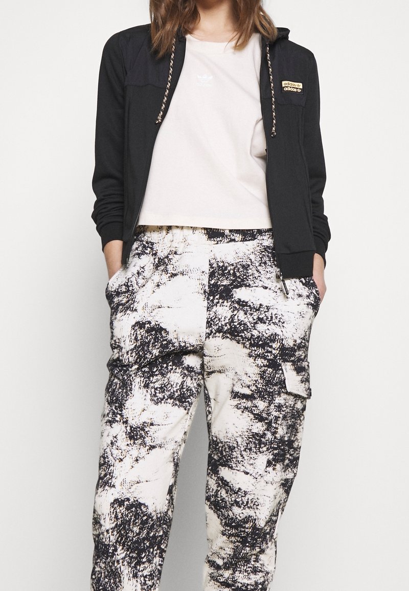 Felpa nera con zip e dettagli dorati, abbinata a una maglietta corta di colore chiaro. Pantaloni stampati in tessuto morbido con motivi astratti in bianco e nero.