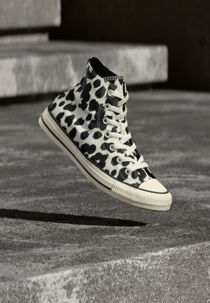 Scarpa da ginnastica alta bianca e nera con motivo leopardato, lacci bianchi e suola in gomma color crema che levita sopra una superficie in cemento.