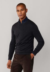 Tmavě šedý svetr s kulatým výstřihem vyrobený z měkkého materiálu, nošený přes navy modrou košili s límečkem. Textura vypadá hladce a má přiléhavý střih.