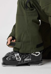 Svart skidpjäxa med silverfärgade beslag, försedd med ett grönt ytterskal. Pjäxan har en slät textur och har en logotyp på sidan.