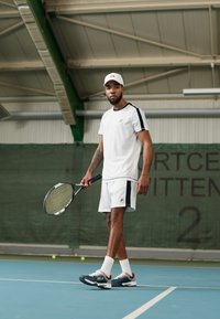 Hvit idretts skjorte med svarte detaljer, matchende shorts og en hvit caps. Holder en tennisracket, iført blå sneakers. Innendørs tennisbane.