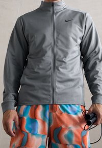 Grå zip-up jakke lavet af glat stof, med høj krave og sort logo. Båret med farverige shorts med et abstrakt mønster.