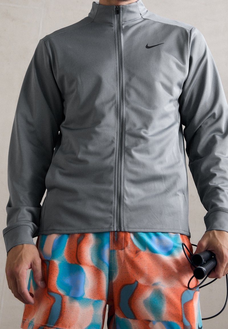Grå zip-up jakke lavet af glat stof, med høj krave og sort logo. Båret med farverige shorts med et abstrakt mønster.