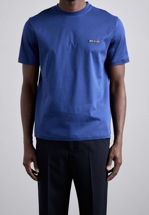 Muž v jednobarevném modrém tričku s krátkým rukávem a malým vícebarevným pruhovaným emblémem na levé horní straně hrudníku a černých kalhotách.