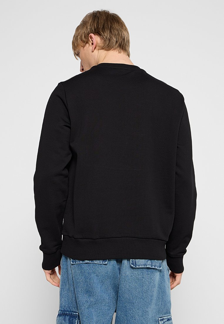 Giovane uomo con capelli corti castano chiaro, che indossa un semplice maglione nero e pantaloni cargo blu in denim, di spalle su uno sfondo bianco.