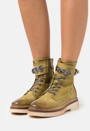 Botas de tobillo de ante verde oliva con herrajes plateados, frontales con cordones, cierre con cremallera y suela texturizada con un borde claro en contraste.