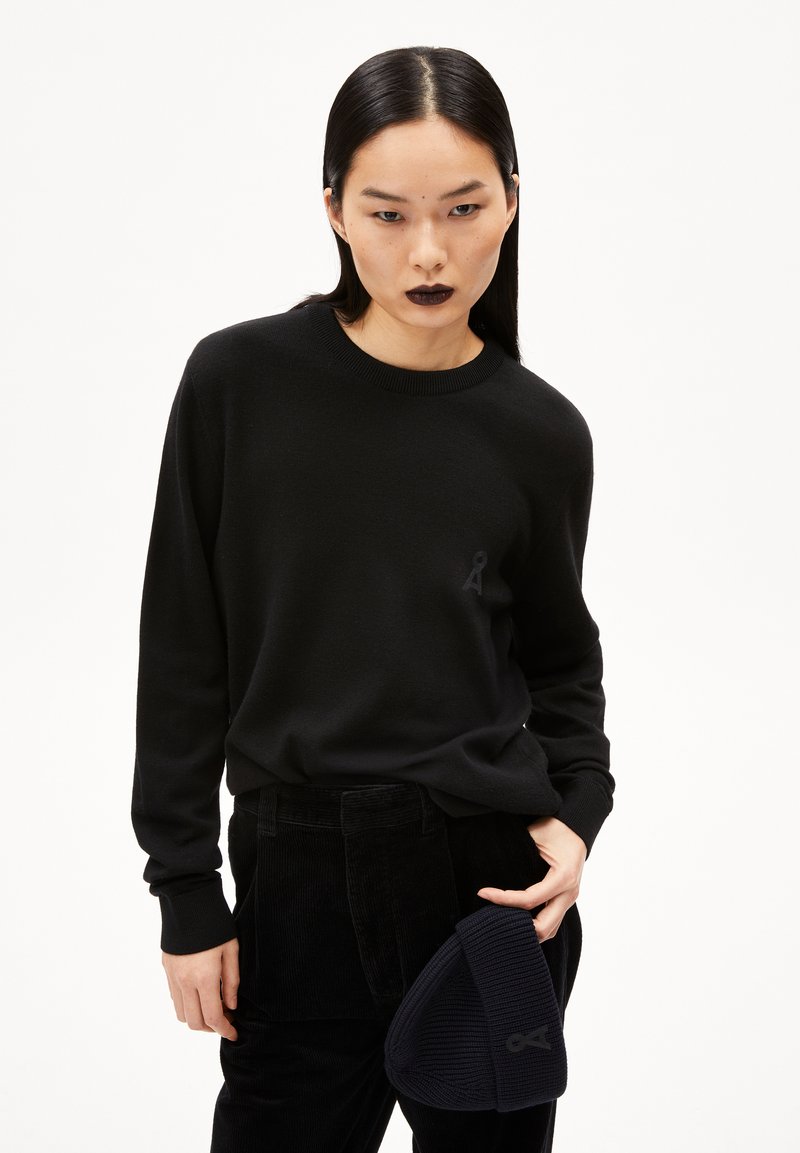 Schwarzer Pullover aus strukturierter Stoff mit Rundhals-Ausschnitt und dezentem Logo. Kombiniert mit schwarzenCordhosen und einer gerippten Mütze.