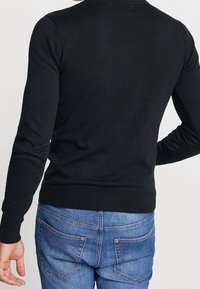 Schwarzer Strickpullover mit langen Ärmeln und rundem Ausschnitt. Verfügt über gerippte Bündchen und Saum sowie ein tailliertes Rückendesign. Wird mit blauer Jeans getragen.