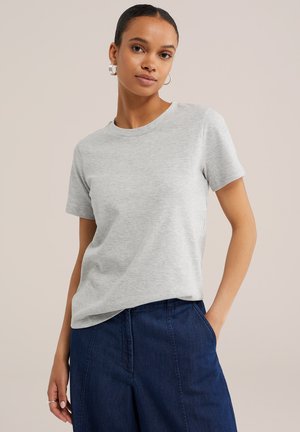 Femeie cu cercei rotunzi, purtând un tricou gri deschis cu mâneci scurte și pantaloni bleumarin cu talie înaltă, stând cu o mână în buzunar.