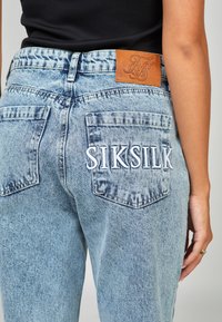 Svijetloplave traperice s teksturiranim finišem, koje imaju kožnu oznaku i izvezeni "SIKSILK" na stražnjem džepu.