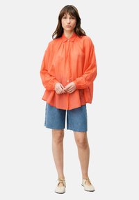 Übergroßes orangefarbenes Hemd mit Faltenlegung, kombiniert mit hellblauen Denim-Shorts und beigen Slippers.