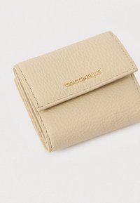Beige leren portefeuille met textuur, gevouwen met het gouden COCCINELLE-logo op de klep, weergegeven tegen een eenvoudige lichte achtergrond.