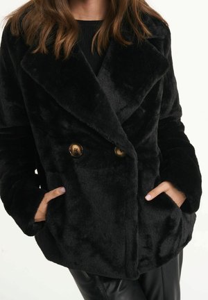 Femme portant un manteau noir en fausse fourrure à double boutonnage avec de grands revers et des boutons, mains dans les poches latérales.