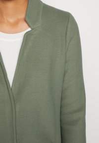 Grön ullblandad kappa med sjalkrage, med en mjuk textur och en öppen design. Buren över en vit ribbad topp.