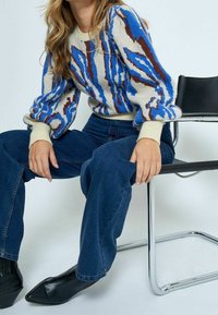 Stickad tröja i kräm, blå och brun abstrakt mönster; ballongsärmar; kombinerad med mörkblå vida jeans och svarta spetsiga stövlar.