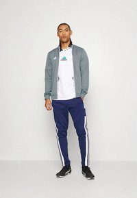 Graue Sportjacke mit weißen und türkisfarbenen Akzenten, getragen über einem weißen T-Shirt mit grünem Logo, kombiniert mit marineblauen Jogginghosen mit weißen Streifen.
