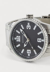 Montre en acier inoxydable argenté avec un cadran noir, de grands marqueurs d'heure blancs et des motifs texturés. Elle dispose d'un guichet de date et d'une couronne texturée.