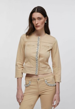 Frau trägt eine beige, verkürzte Jacke und Hose mit silbernem Paillettenbesatz, steht vor einem schlichten hellgrauen Hintergrund.