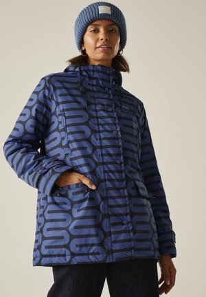 Kobieta w niebieskiej, wzorzystej zimowej kurtce i niebieskiej dzianinowej czapce, z jedną ręką w kieszeni kurtki, na neutralnym tle.