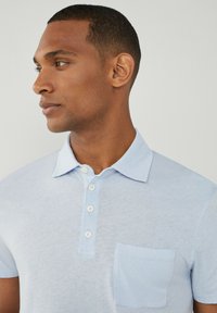 Svetlo modrá polokošela z jemnej látky, s gombíkovým zapínaním, golierom a jedným vreckom na hrudi. Hladká textúra a ležérny dizajn.