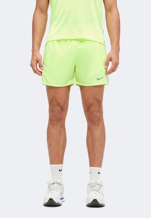 Homme portant un short Nike jaune néon, des chaussettes Nike blanches et des chaussures de sport blanches, debout devant un fond clair uni.