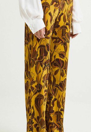 Pantalon à jambes larges en tissu jaune moutarde, avec un motif floral marron et rouge. Texture lisse, porté avec un chemisier blanc.