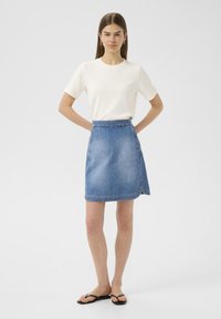 Jupe en denim bleu clair, avec une taille ajustée, une forme en A et un ourlet brut. Associée à un haut blanc à manches courtes et des tongs noires.