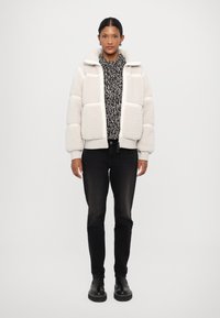 Puffer takki pehmeästä off-white materiaalista, jossa on tikattuja osia, korkea kaulus ja etuvetoketju, yhdistettynä tummiin farkkuihin ja mustiin saappaisiin.