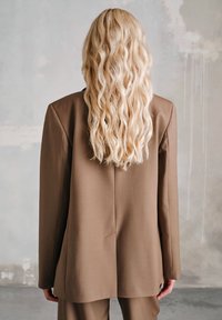 Brązowa, dopasowana marynarka o strukturze, z prostą sylwetką z tyłu i gładką teksturą. Widoczne długie, falowane włosy.