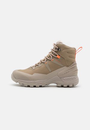 Beige wandelschoen met een bovenwerk van gestructureerde stof, rubberen zool en oranje accenten. Heeft hoge enkelsupport en een versterkte neuskap.