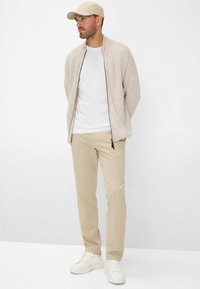 Lichtbeige gebreide cardigan met een rits aan de voorkant, een wit T-shirt, beige broeken en witte sneakers. De pet is lichtbeige met een blauwe accent.