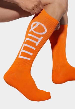 Osoba podciągająca jasno-pomarańczowe, sięgające do kolan skarpetki z białym logo na jednej nodze na jednolitym tle.