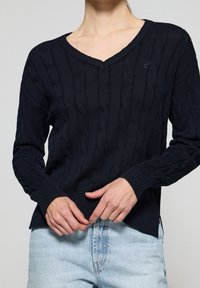 Pull en maille marine avec un col en V, présentant des motifs en relief et des poignets côtelés. Porté par-dessus un jean bleu clair, montrant une coupe décontractée et une texture.